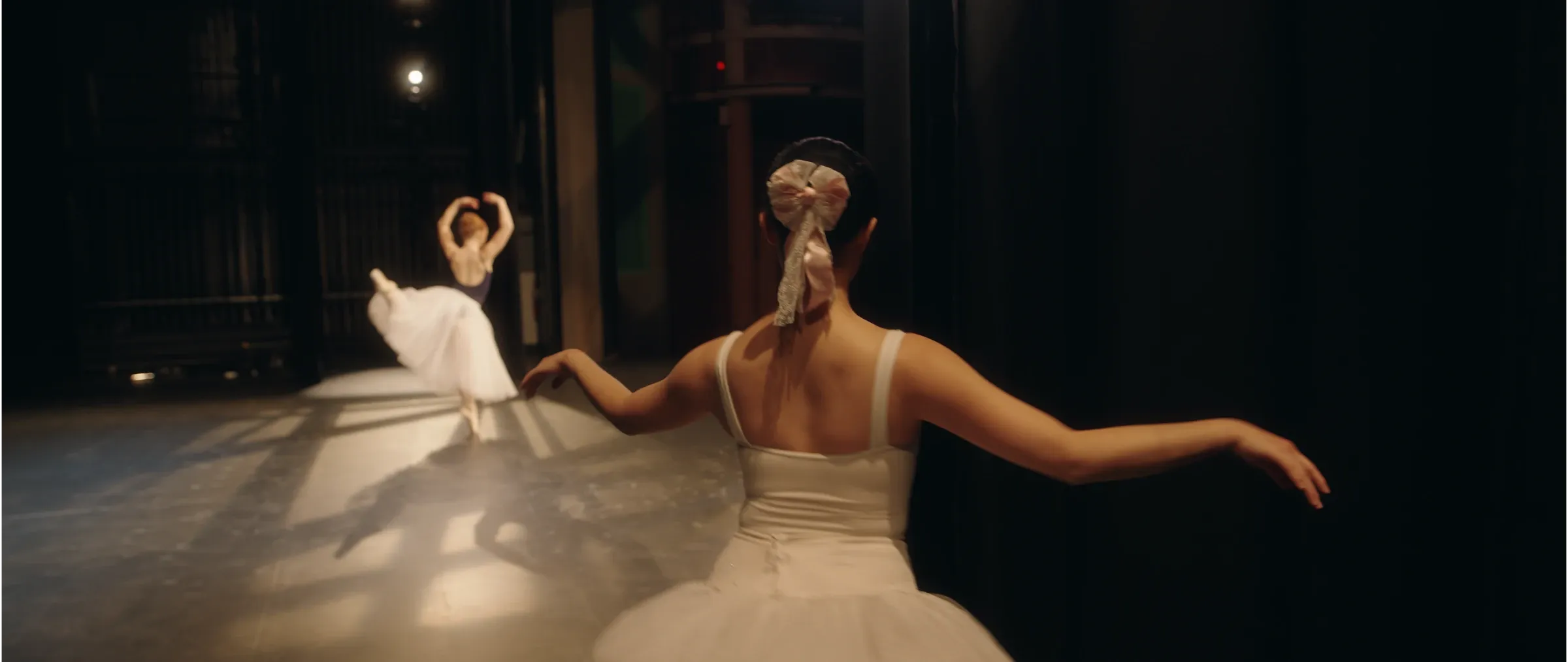 Two ballerinas perform on a dark stage.